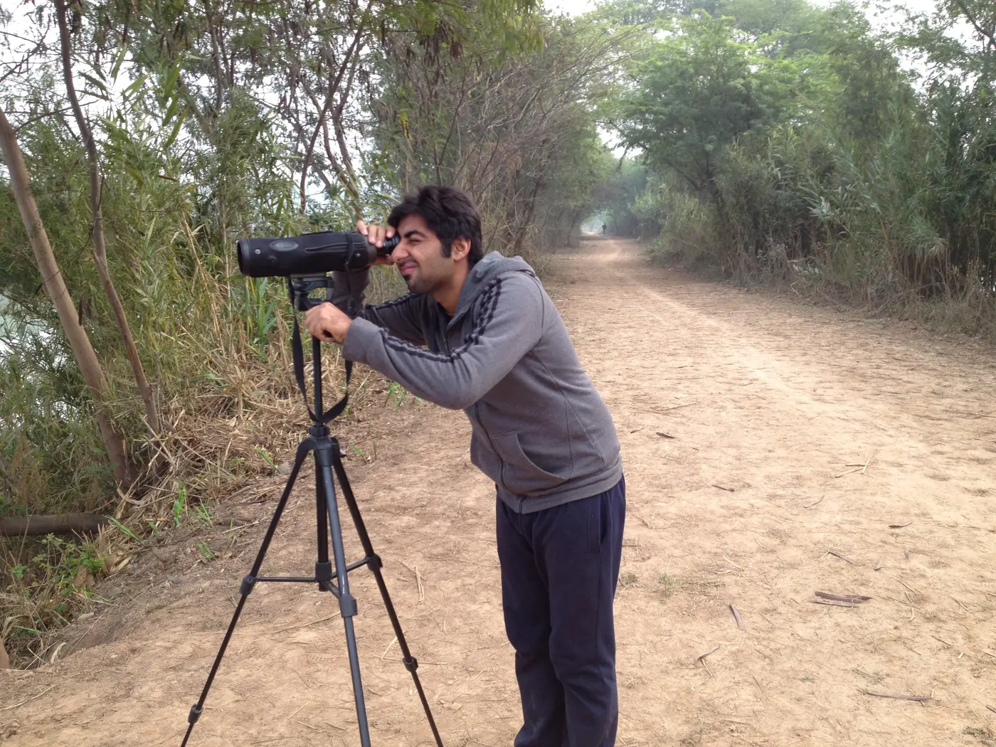 A visitor birdwatching with a spotting scope along a tree-lined dirt path in Chandigarh's forest cover, the kind of quiet walking trail found in the Botanical Garden