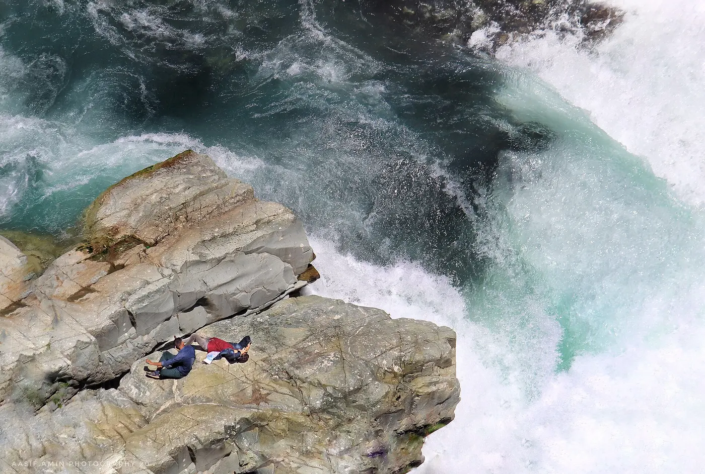 Visitors seated on wet rocks right at the edge of Aharbal Falls, illustrating how close and slippery the unrailed sections can be