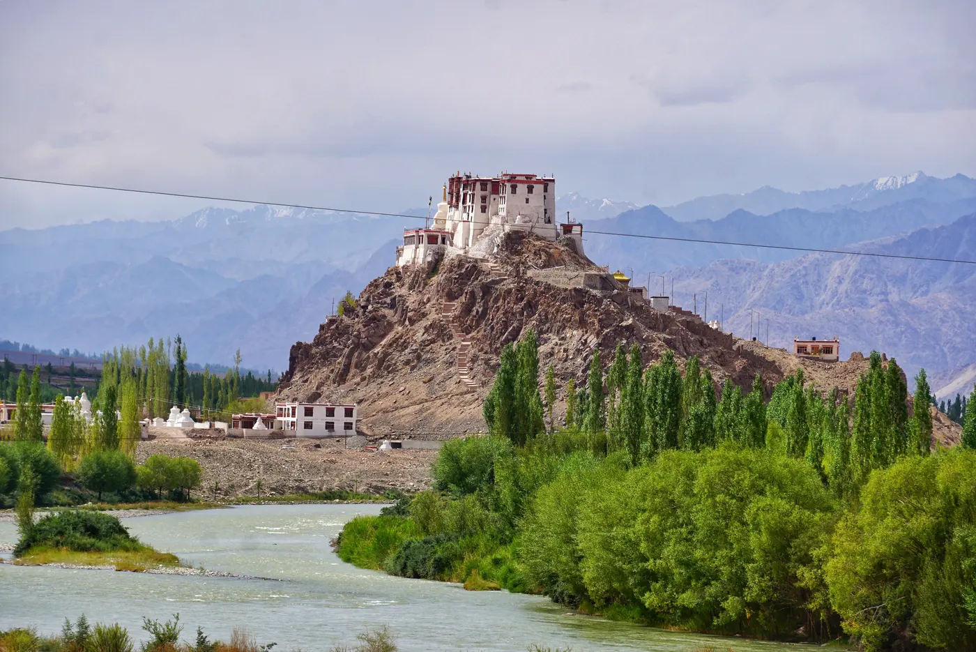 Stakna Monastery perched on a rocky outcrop above the Indus River, framed by poplar trees and distant Himalayan peaks