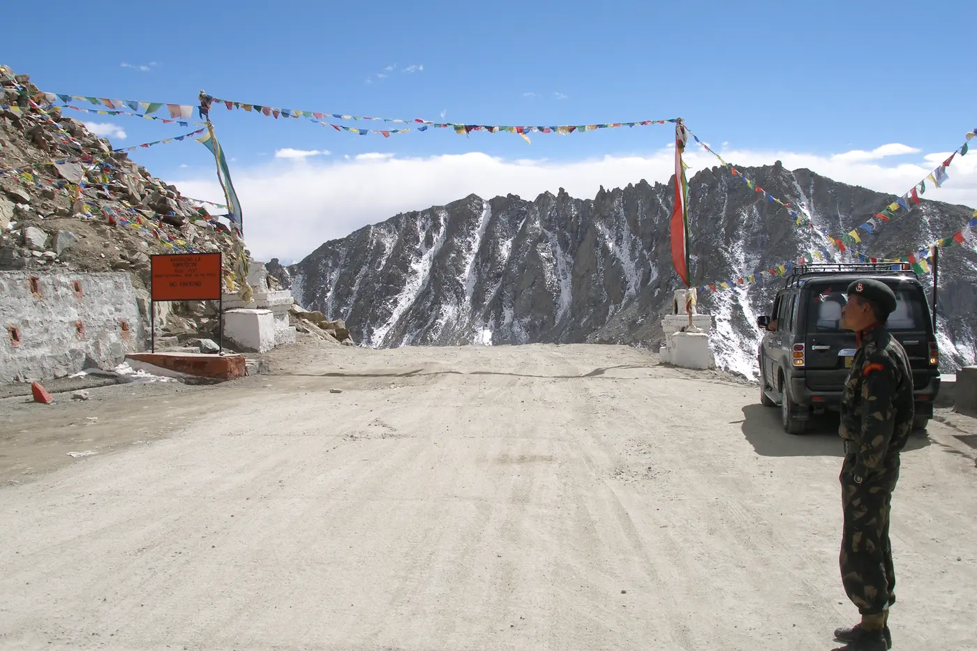 Army checkpost near Khardung La with prayer flags strung overhead, a parked jeep, and a soldier on duty