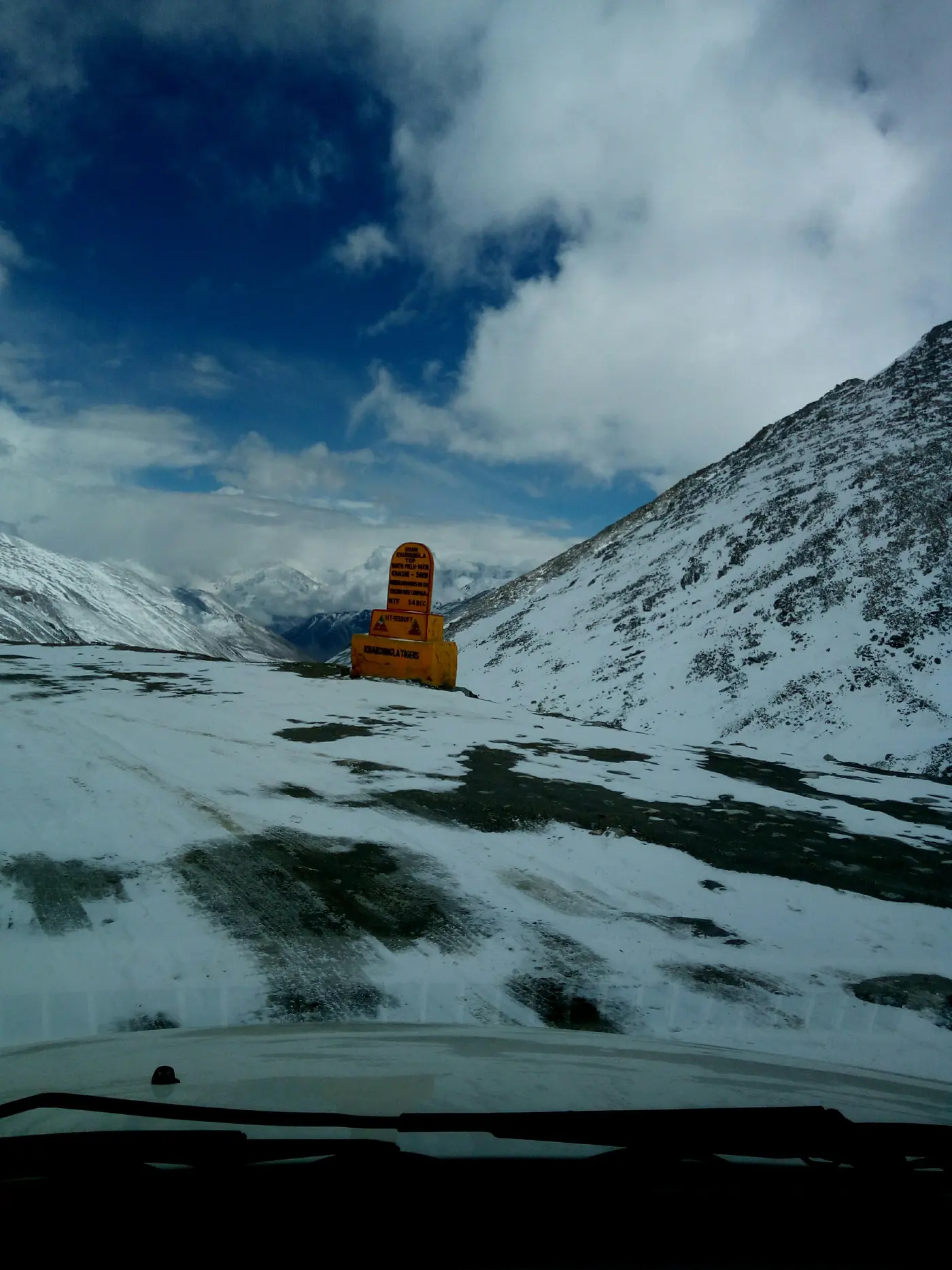 The yellow Khardung La summit marker signboard listing distances, surrounded by snow at the top of the pass