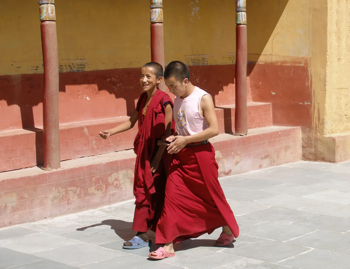 Two young monks in maroon robes walking through a sunlit courtyard at Thiksey Monastery, Ladakh