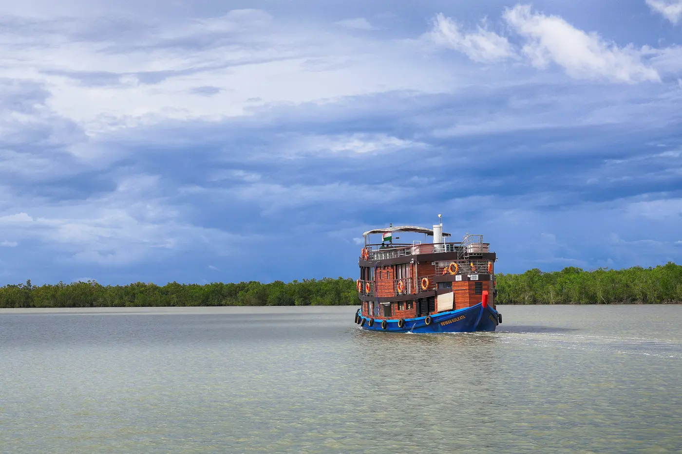 A tourist boat cruising a wide tidal creek lined with mangrove forest in the Sundarbans
